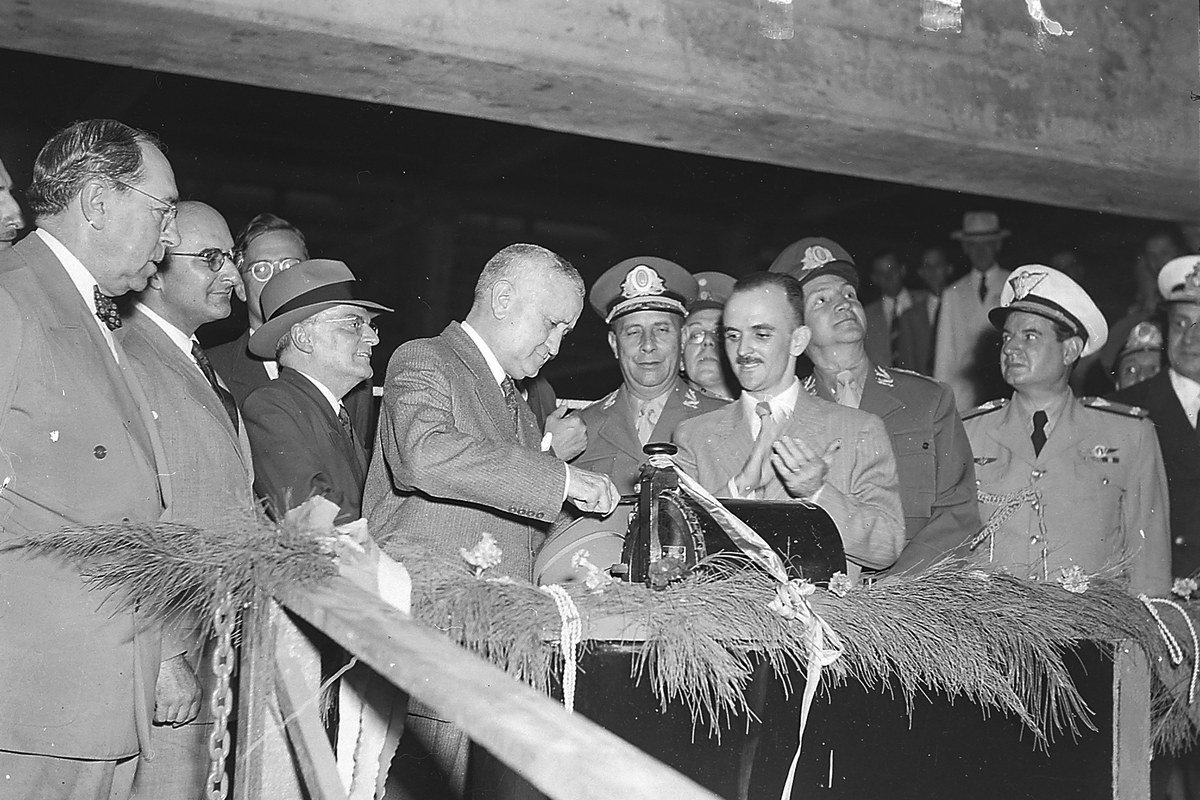 Foto histórica em preto e branco da cerimônia de inauguração da Companhia Siderúrgica Nacional (CSN) em 1946, com o presidente Eurico Gaspar Dutra acionando o botão simbólico ao lado de autoridades e militares
