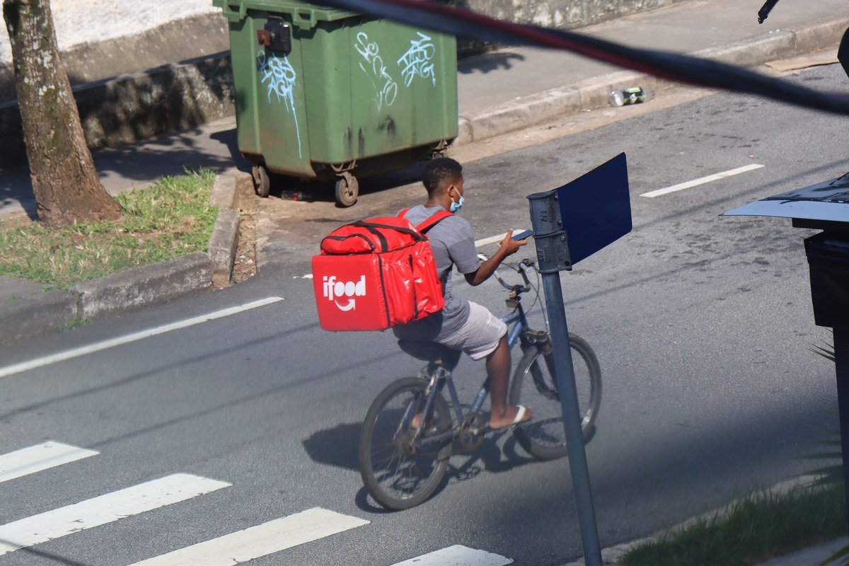 Entregador de bicicleta com bag térmica vermelha do iFood e máscara de proteção durante a pandemia, atravessando rua com faixa de pedestre