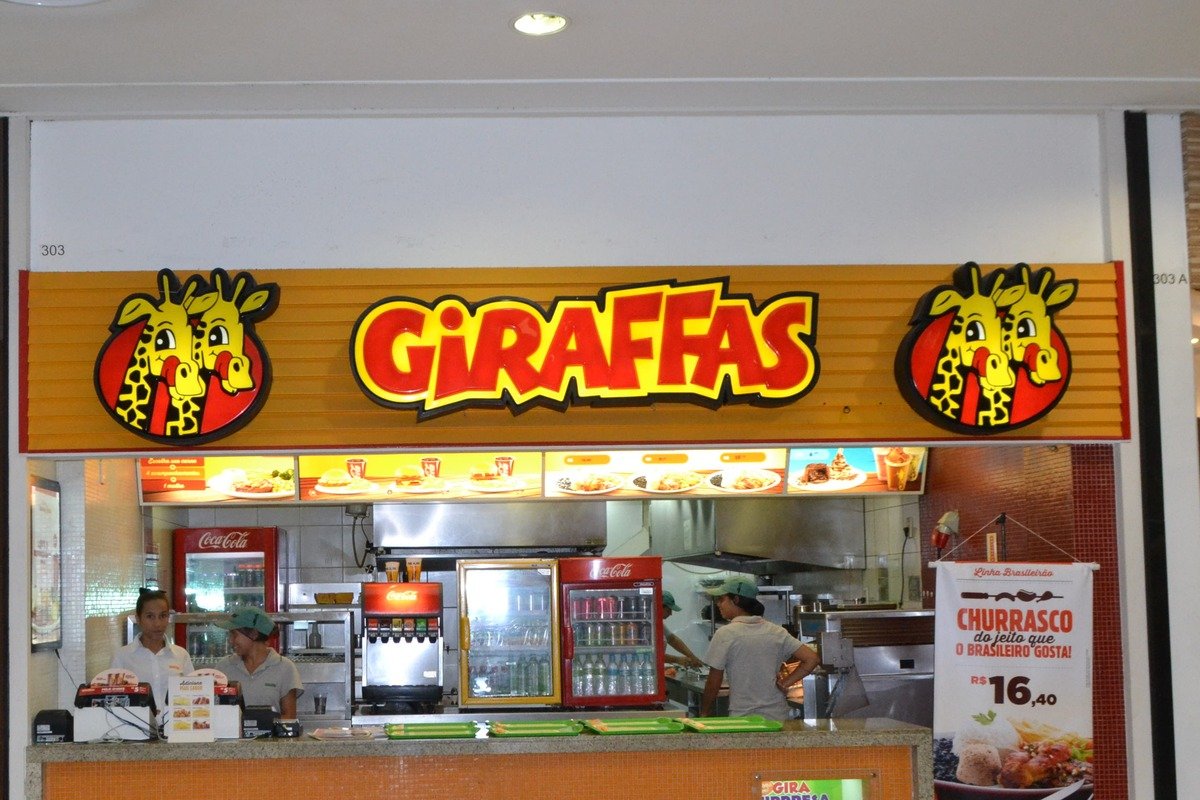 Balcão de atendimento de uma loja Giraffas em shopping, com o letreiro da marca, painéis de cardápio mostrando pratos prontos e funcionários uniformizados trabalhando