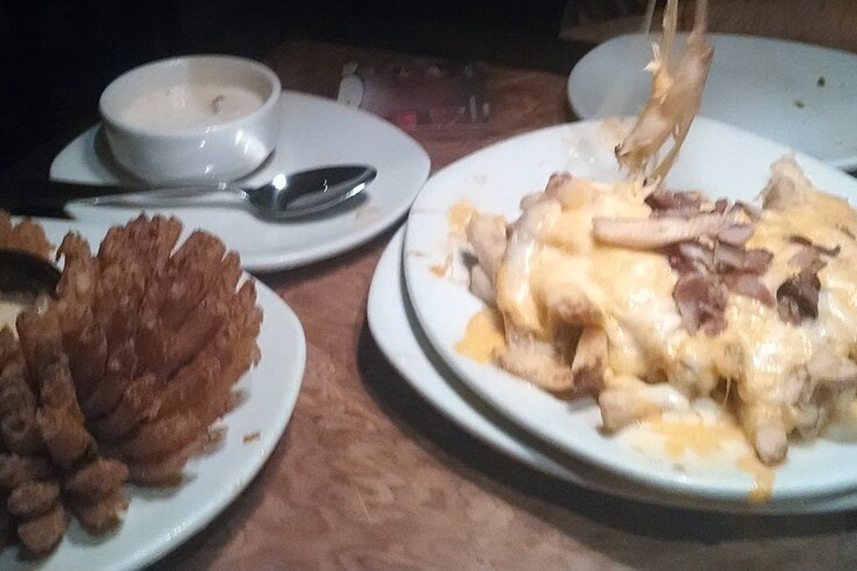 Prato de Bloomin' Onion, cebola cortada em pétalas e frita, ao lado de porção de batatas com queijo e bacon em uma mesa de restaurante Outback