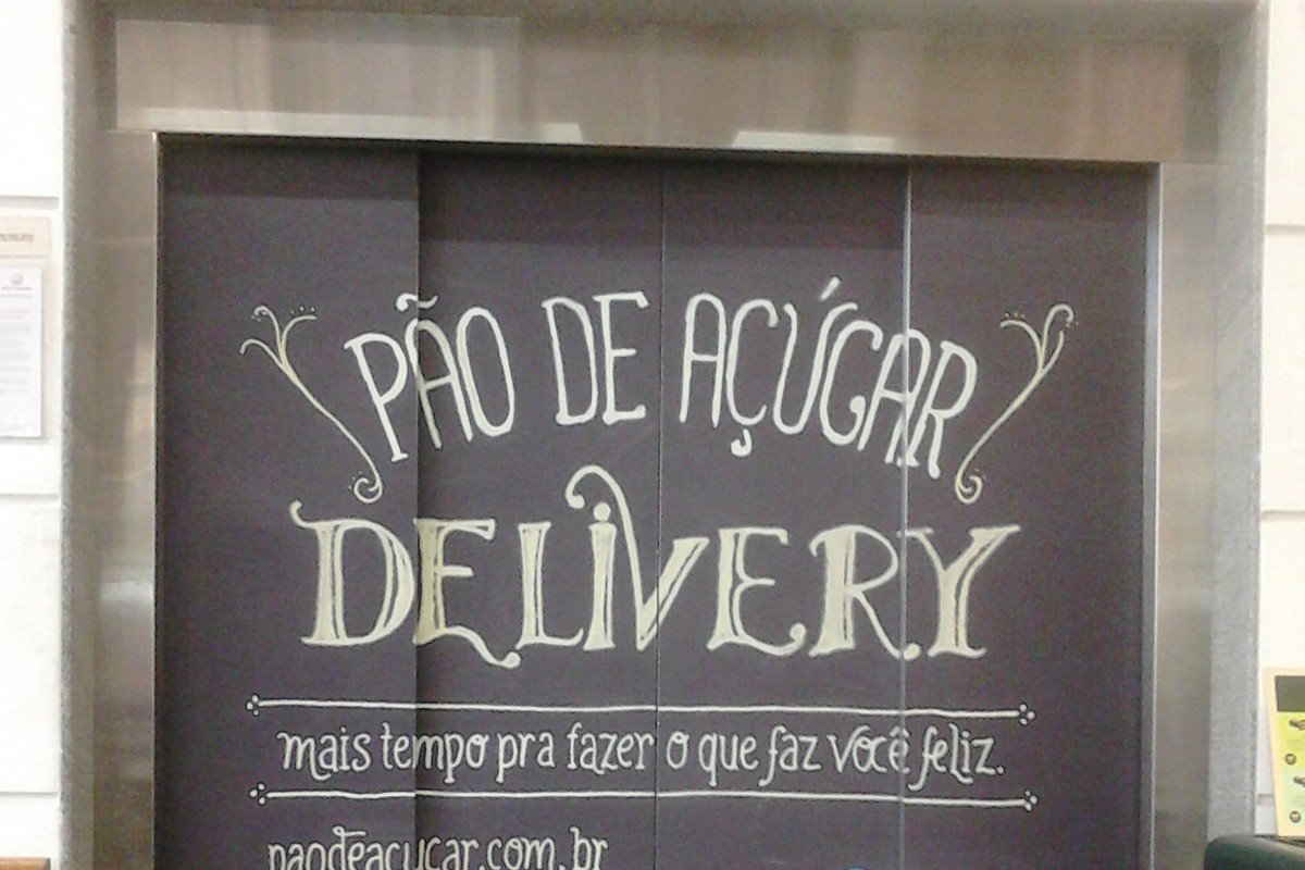 Identidade visual da marca Pão de Açúcar em material de divulgação de serviço de entrega Painel de elevador decorado com a propaganda 'Pão de Açúcar Delivery', em letras estilizadas sobre fundo escuro, com um carrinho de compras em primeiro plano