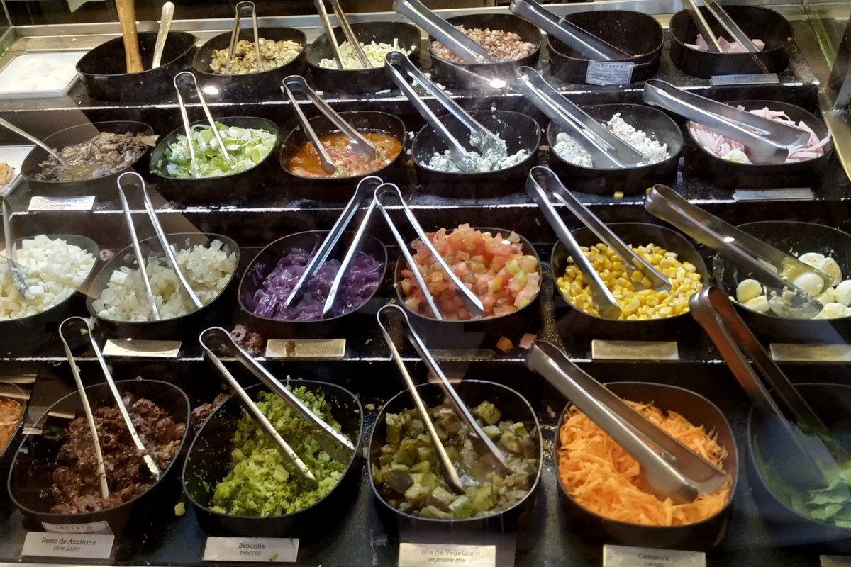 Balcão de vidro com dezenas de potes de ingredientes para massa em uma loja Spoleto, incluindo legumes picados, molhos e queijos, com pinças de servir sobre cada pote
