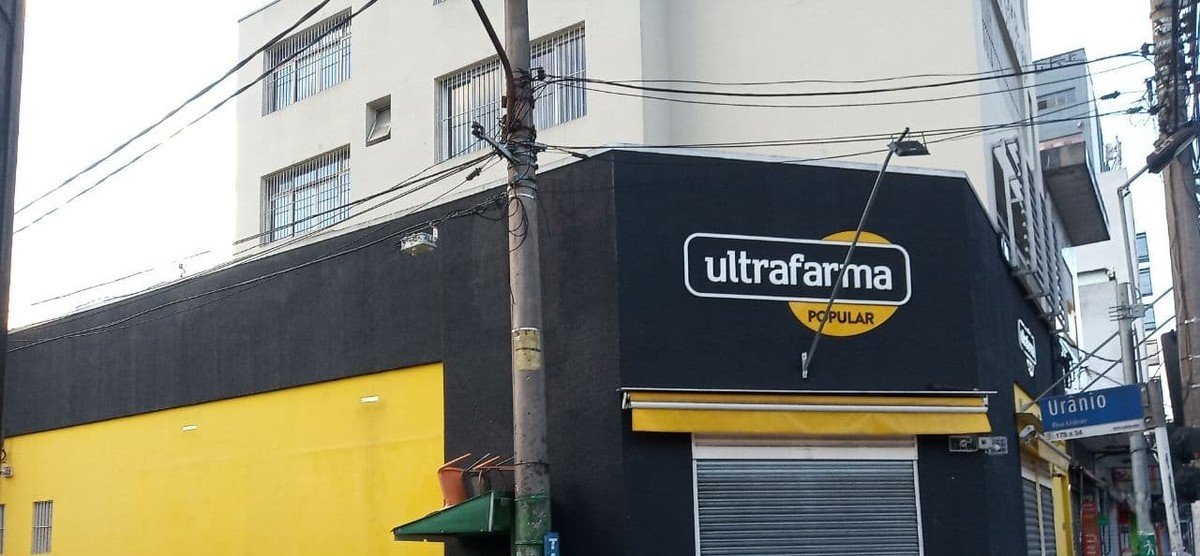 Fachada de loja com letreiro oval amarelo e preto escrito Ultrafarma Popular, em esquina de rua