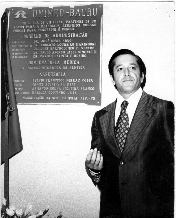 Retrato de Edmundo Castilho, fundador da Unimed, em traje social, ao lado de uma placa comemorativa da inauguração da sede própria da Unimed-Bauru em fevereiro de 1978
