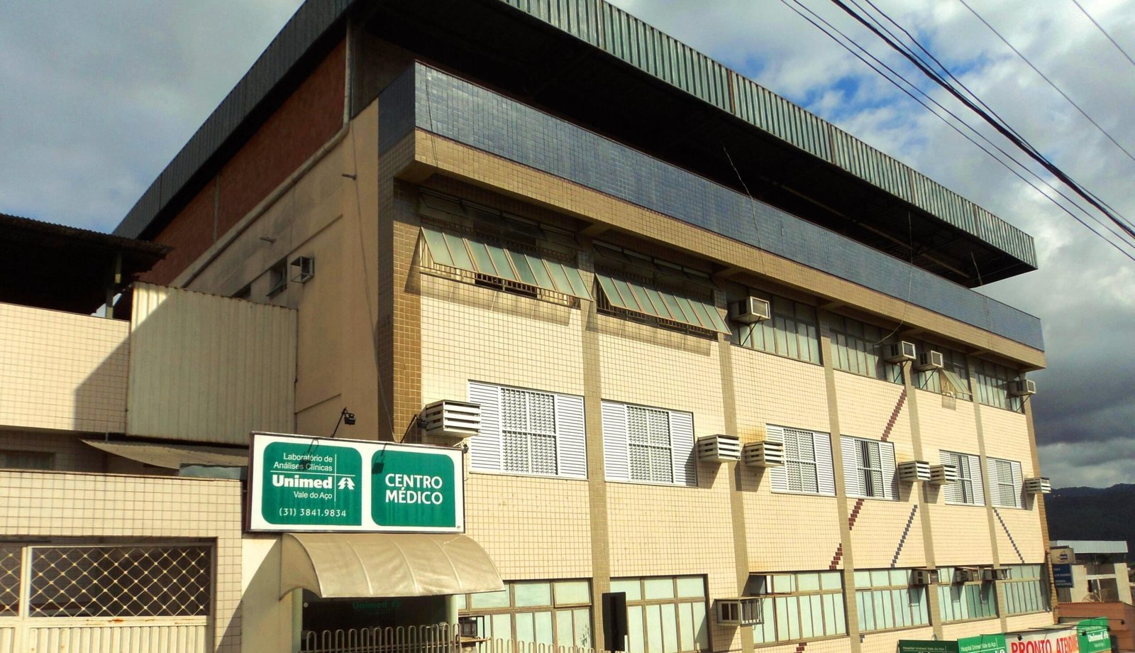 Fachada de um prédio de tijolos e azulejos bege com uma placa verde escrito 'Laboratório de Análises Clínicas Unimed Vale do Aço' e 'Centro Médico', em Coronel Fabriciano, Minas Gerais