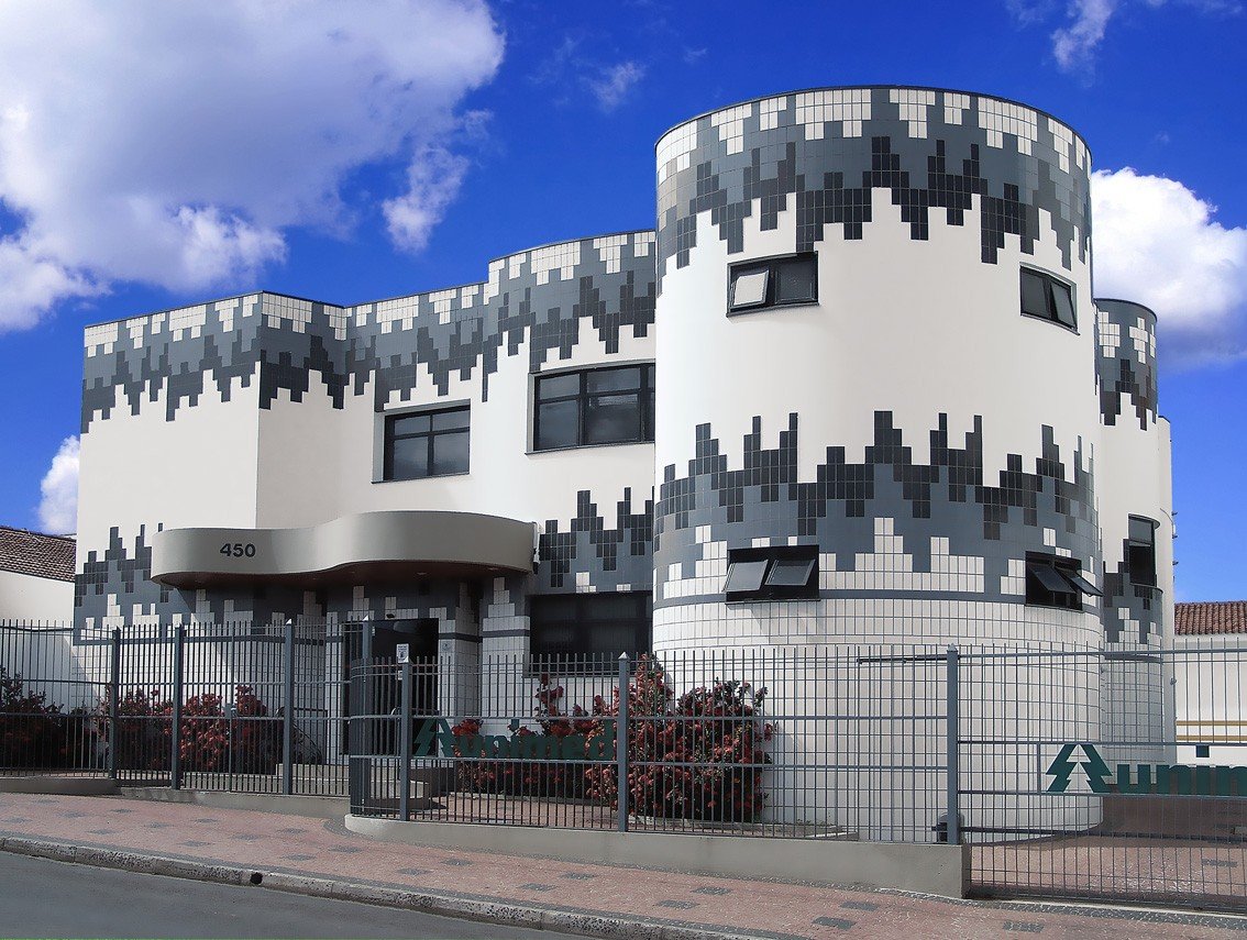 Fachada moderna de um prédio de dois andares com revestimento em mosaico geométrico branco, cinza e preto, cercado por grades e plantas, com o logo da Unimed visível no portão