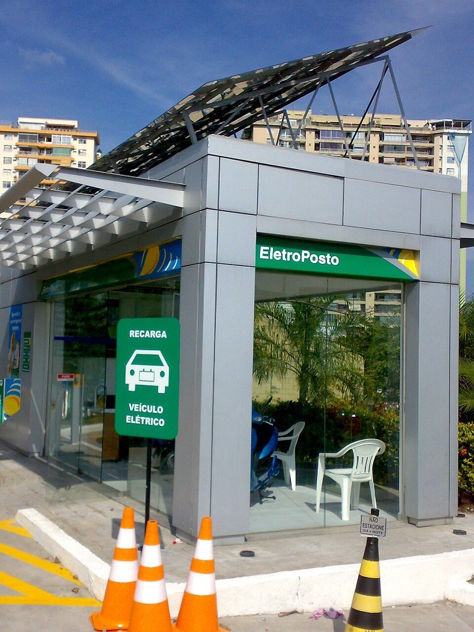 O primeiro eletroposto do Brasil, instalado em 2009, mais de uma década antes do boom dos carros elétricos Estrutura moderna de vidro e metal com painéis solares no teto e a placa 'EletroPosto' e 'Recarga Veículo Elétrico', sem carros nem pessoas visíveis