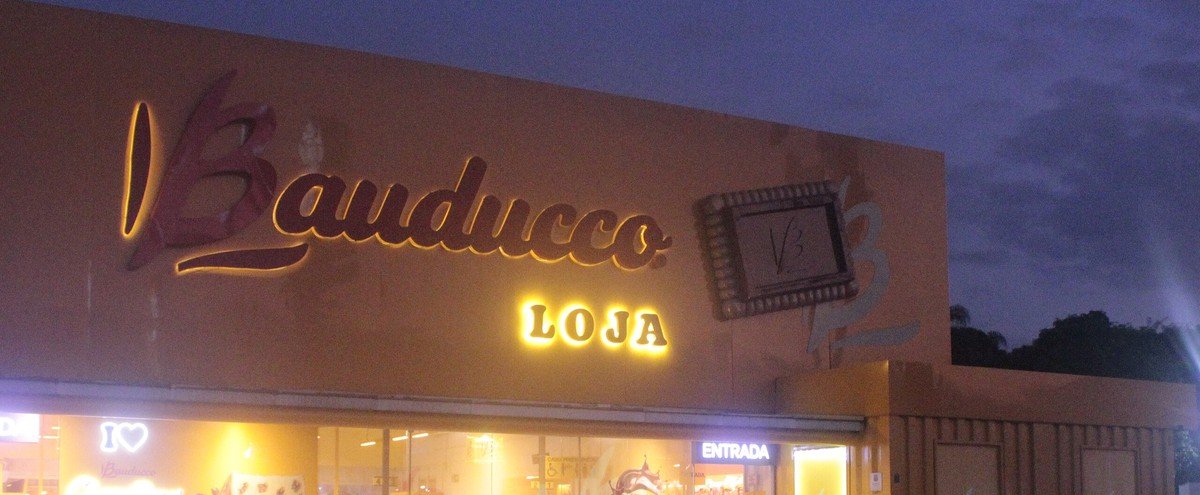 Fachada iluminada de uma loja Bauducco, com o logotipo da marca e a palavra loja, à beira da Via Dutra em Roseira (SP)