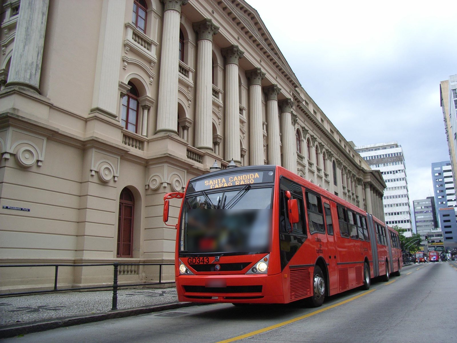 Ônibus biarticulado vermelho com chassi Volvo B12M passando em frente ao prédio histórico da Universidade Federal do Paraná, em Curitiba