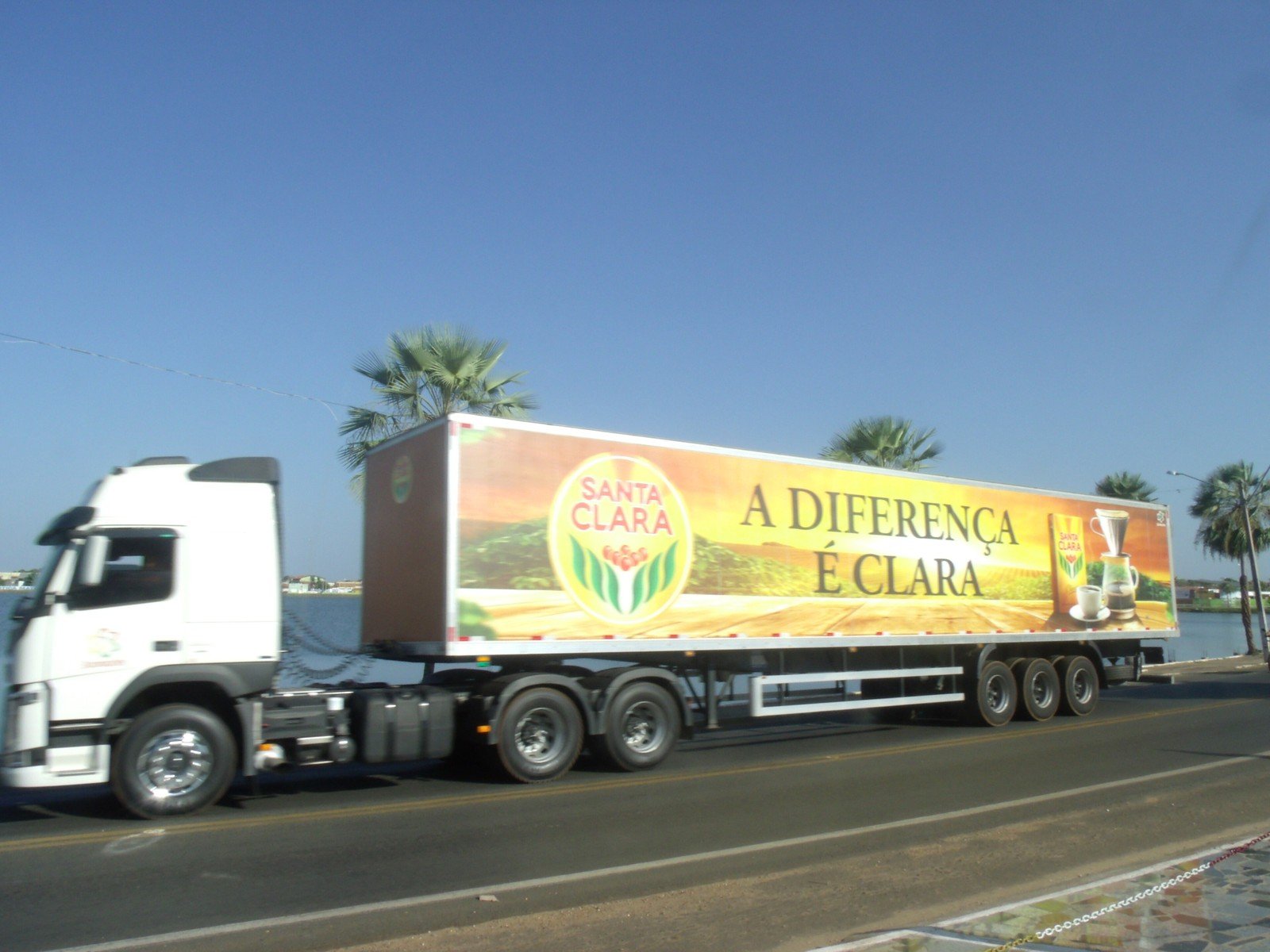 Carreta com propaganda do Café Santa Clara, marca do Grupo 3corações, trafegando em uma rodovia à beira de um lago
