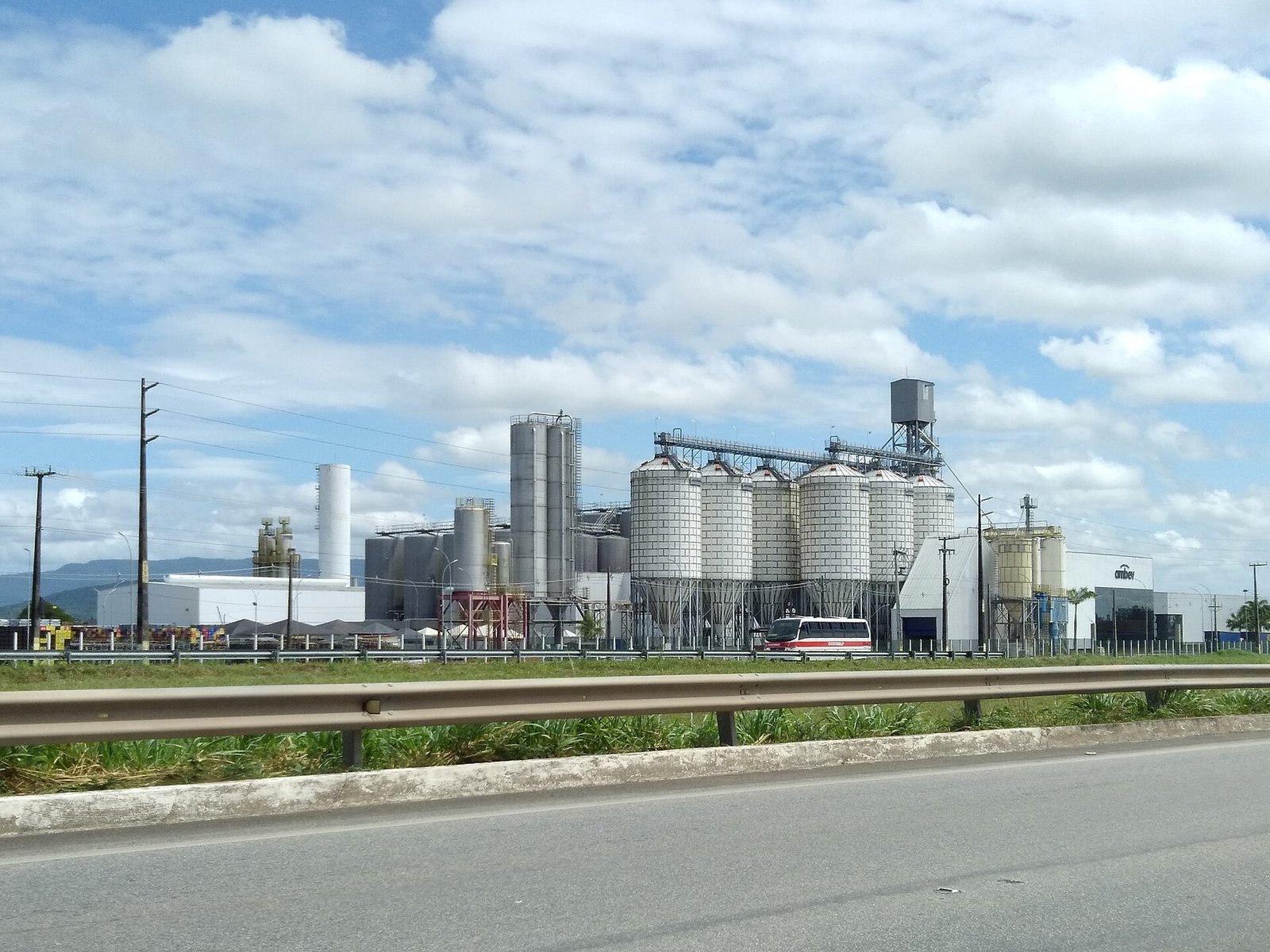 Silos e tanques metálicos de uma fábrica de cerveja da Ambev à beira de uma rodovia, sob céu azul com nuvens