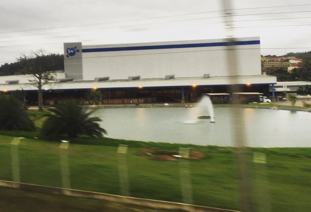 Quem é o dono da Ypê? A fabricante de Amparo pertence à família Beira 2 Galpão branco da fábrica da Ypê com o logotipo azul da marca na fachada, atrás de um lago com chafariz e um gramado