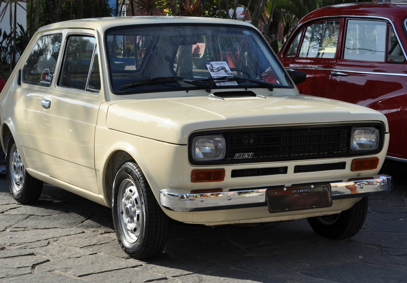 Fiat 147 bege, do primeiro modelo, estacionado em uma praça com calçamento de pedras, ao lado de um carro antigo vermelho