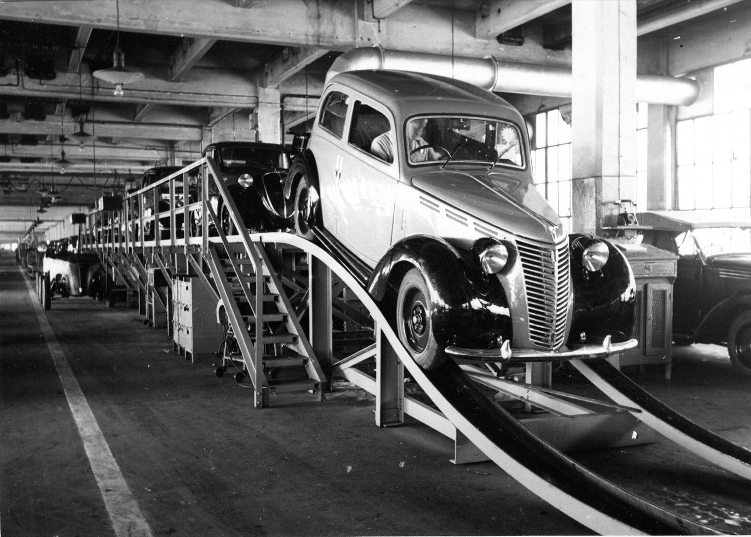 Foto em preto e branco de uma linha de montagem antiga, com carros Fiat 1100 subindo por uma rampa entre estruturas metálicas