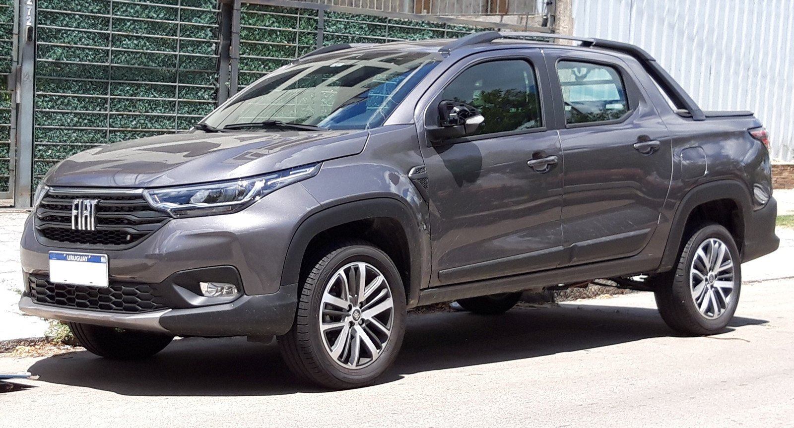 Picape Fiat Strada cinza, cabine dupla, estacionada em uma rua ao lado de um portão coberto de plantas