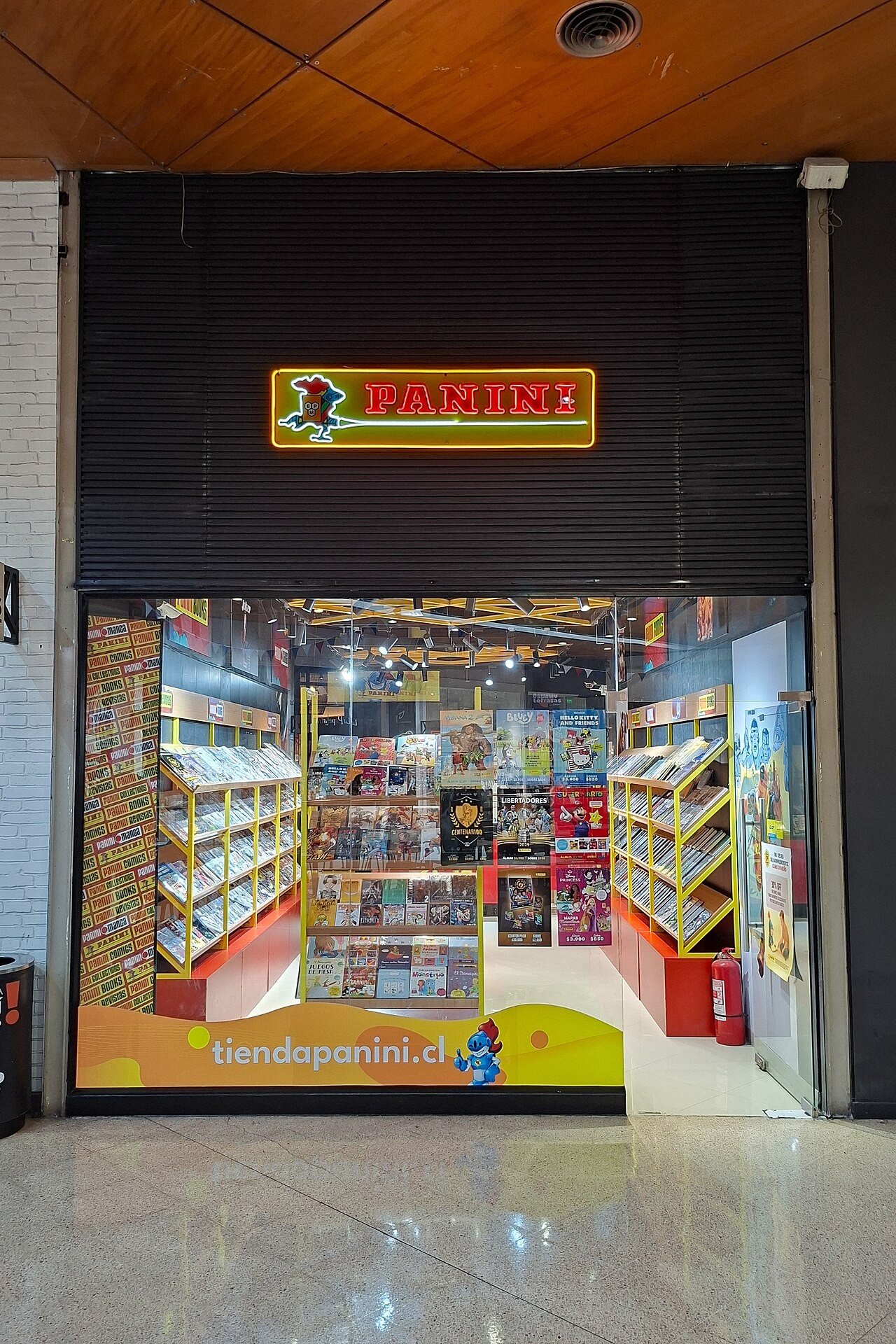 Fachada de uma loja da Panini em shopping, com o letreiro da marca em neon e prateleiras de álbuns e revistas