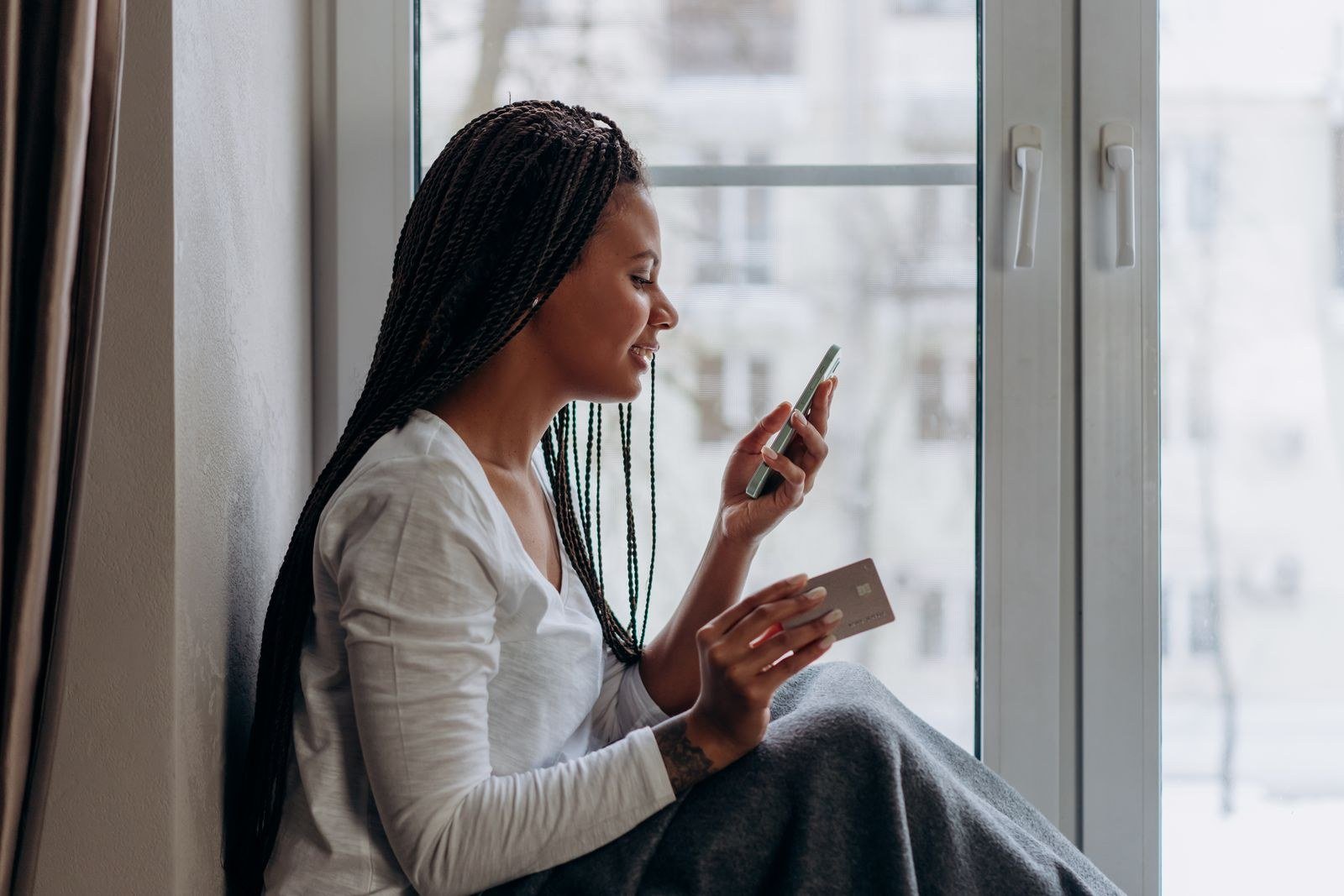 Melhor dia de compra no cartão: como descobrir e ganhar mais prazo para pagar 5 Mulher sentada perto da janela segurando um cartão de crédito e o celular