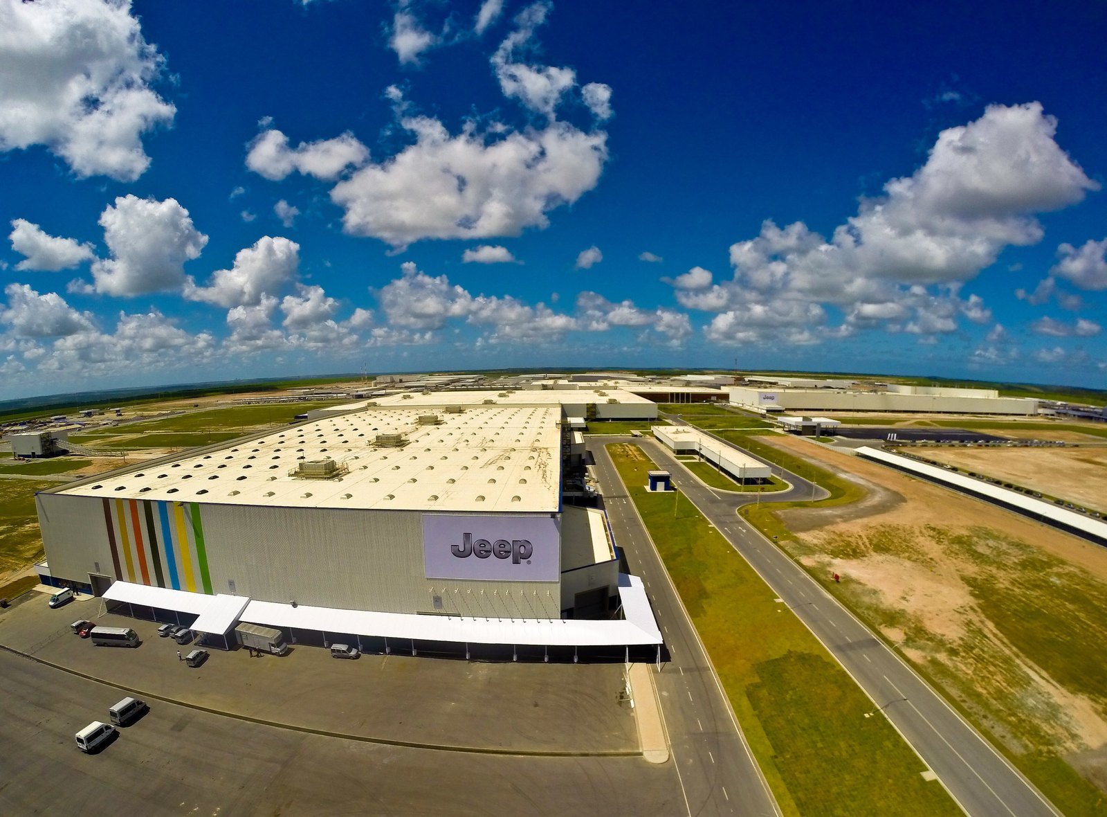 Vista aérea de uma grande fábrica com o letreiro da Jeep na fachada, cercada por pistas e terrenos, sob céu azul com nuvens